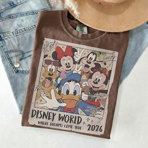 May include: Brown t-shirt featuring a Disney World graphic with Mickey Mouse, Minnie Mouse, Donald Duck, Pluto, and Goofy. The graphic includes the text "Disney World, Where Dreams Come True 2026". The shirt is paired with blue jeans and a tan hat.