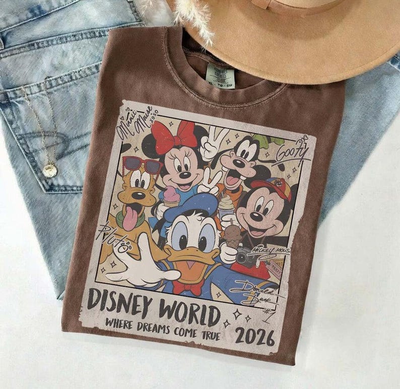 May include: Brown t-shirt featuring a Disney World graphic with Mickey Mouse, Minnie Mouse, Donald Duck, Pluto, and Goofy. The graphic includes the text "Disney World, Where Dreams Come True 2026". The shirt is paired with blue jeans and a tan hat.