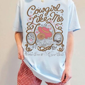 May include: A light blue t-shirt featuring a graphic print with the text Cowgirl Like Me, Takes One to Know One, and Eyes full of Stars. The design includes a rope border, a pink cowboy hat, a horseshoe, musical notes, and small star accents.