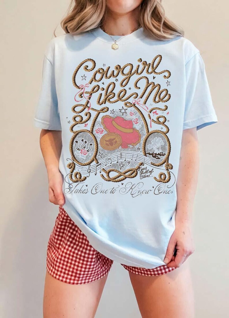 May include: A light blue t-shirt featuring a graphic print with the text Cowgirl Like Me, Takes One to Know One, and Eyes full of Stars. The design includes a rope border, a pink cowboy hat, a horseshoe, musical notes, and small star accents.