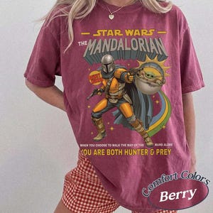 May include: A berry-colored t-shirt featuring a graphic of The Mandalorian and Grogu. The text reads STAR WARS THE MANDALORIAN, WHEN YOU CHOOSE TO WALK THE WAY OF THE MANDALORE YOU ARE BOTH HUNTER & PREY. The design includes a sunburst and stars.