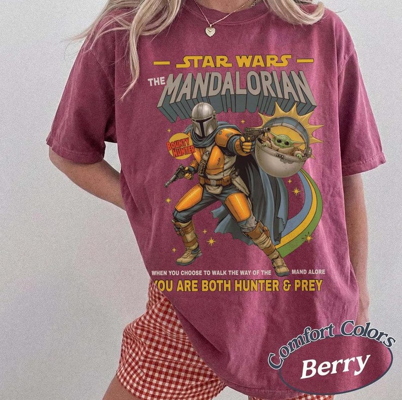 May include: A berry-colored t-shirt featuring a graphic of The Mandalorian and Grogu. The text reads STAR WARS THE MANDALORIAN, WHEN YOU CHOOSE TO WALK THE WAY OF THE MANDALORE YOU ARE BOTH HUNTER & PREY. The design includes a sunburst and stars.
