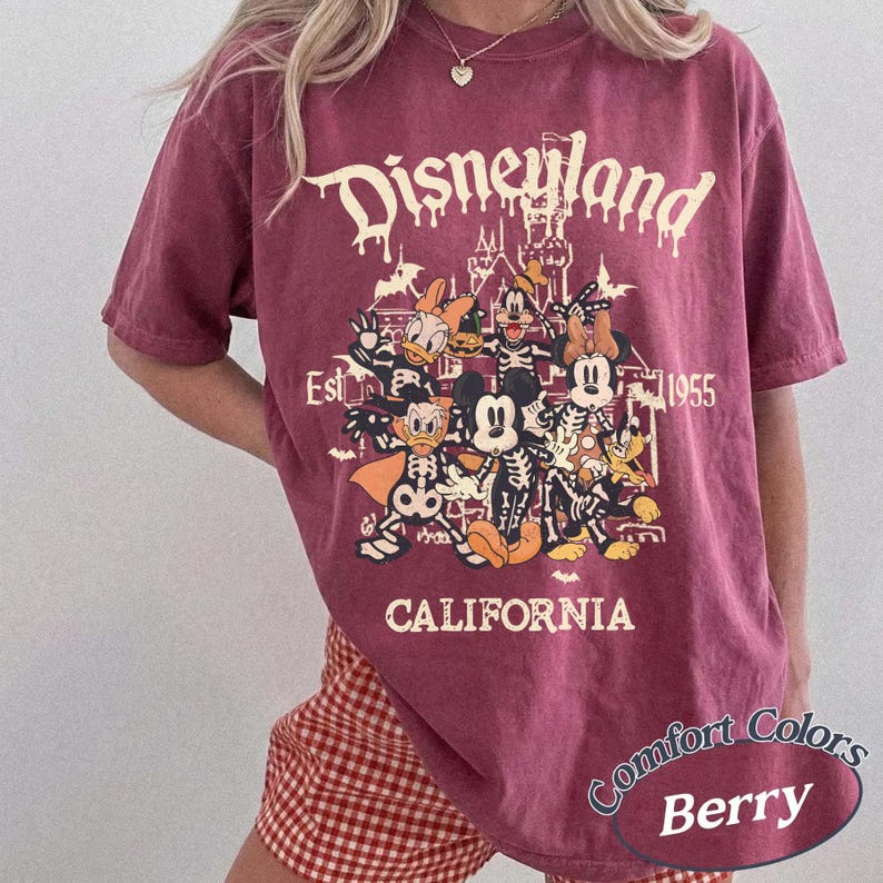 May include: Berry-colored t-shirt with a Disneyland graphic featuring Disney characters in skeleton costumes. The text "Disneyland California Est. 1955" is printed above the graphic. The shirt is a Comfort Colors brand.