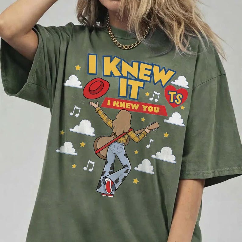 May include: An olive green t-shirt featuring a graphic of a person playing an acoustic guitar. The text reads I KNEW IT and I KNEW YOU. The design includes a red hat, a red heart with TS, white clouds, yellow stars, and musical notes.