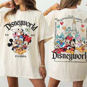 May include: Cream-colored t-shirt featuring the text "Disneyworld" with a graphic of Disney characters. The front of the shirt displays Mickey, Minnie, Donald, Daisy, Goofy, and Pluto. The back shows the Disney castle and more characters.