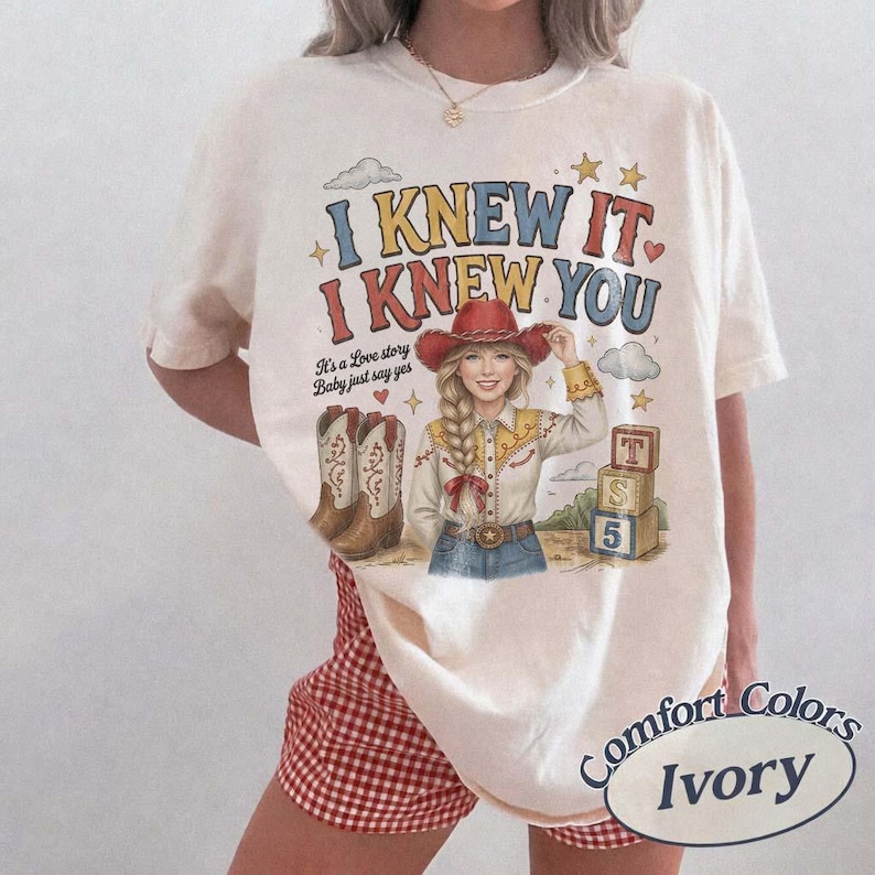 May include: An ivory t-shirt featuring a graphic print with the text I KNEW IT I KNEW YOU and It's a Love story Baby just say yes. The design includes a blonde person in a red cowboy hat, a pair of cowboy boots, and a stack of three alphabet blocks.