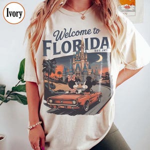 May include: Ivory-colored t-shirt with a graphic that says "Welcome to Florida" and "Est. 1971." The graphic features Mickey and Minnie Mouse in a red convertible in front of a castle and palm trees.