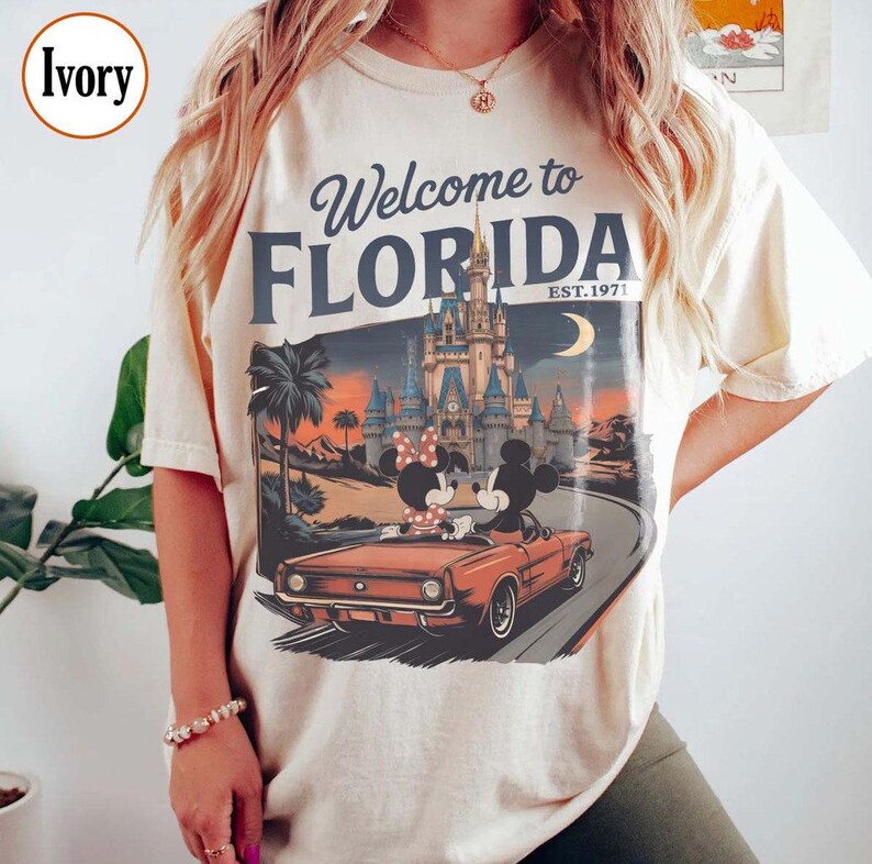 May include: Ivory-colored t-shirt with a graphic that says "Welcome to Florida" and "Est. 1971." The graphic features Mickey and Minnie Mouse in a red convertible in front of a castle and palm trees.