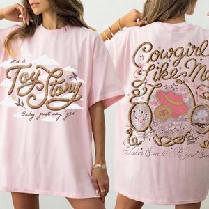 May include: A light pink t-shirt featuring a front graphic with the text "It's a Toy Story" in rope-style lettering and clouds. The back graphic displays the text "Cowgirl Like Me" and "Takes One to Know One" with rope borders, a pink hat, and musical notes.