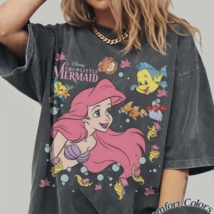 May include: Dark gray t-shirt featuring a graphic of Ariel from Disney's The Little Mermaid. Ariel has pink hair and a purple seashell bra. The shirt also features the text "The Little Mermaid" and various sea creatures. The shirt is labeled "Comfort Colors Pepper".