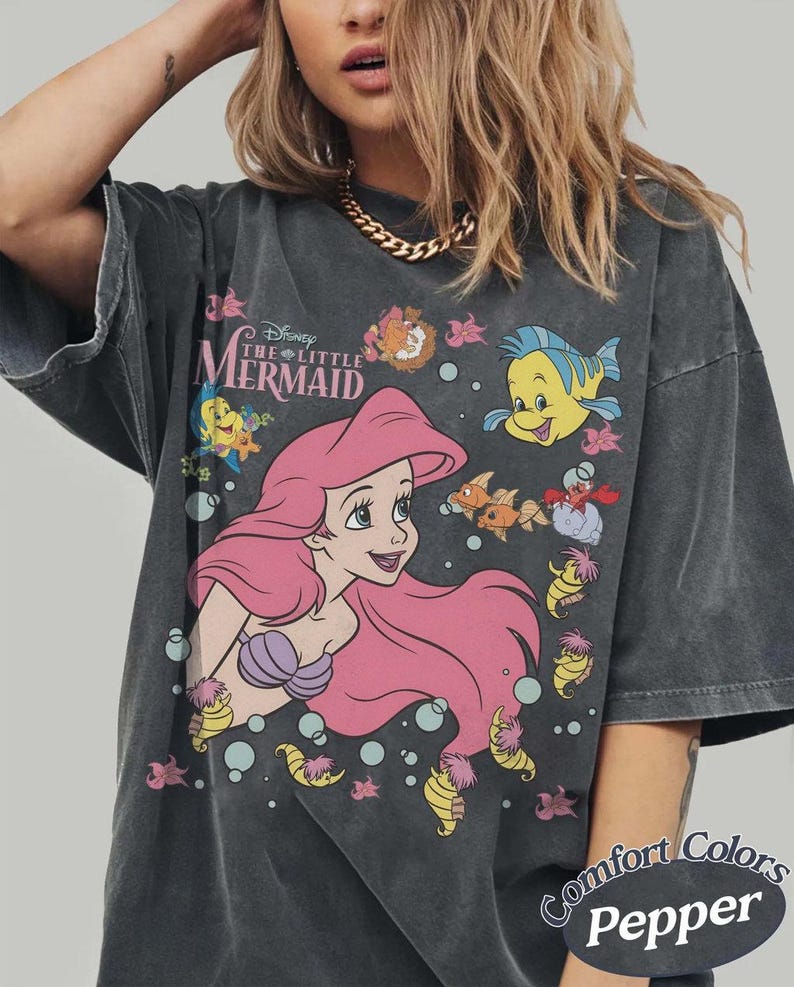 May include: Dark gray t-shirt featuring a graphic of Ariel from Disney's The Little Mermaid. Ariel has pink hair and a purple seashell bra. The shirt also features the text "The Little Mermaid" and various sea creatures. The shirt is labeled "Comfort Colors Pepper".