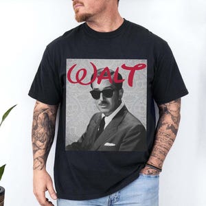 May include: Black t-shirt featuring a black and white image of a man wearing sunglasses and a suit. The word "WALT" is written in red cursive above the image. The shirt is a classic crew neck.