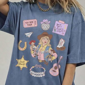 May include: A blue t-shirt featuring a graphic print of a person in a cowboy hat, surrounded by illustrations including a Toy Box Tour ticket, a disco ball, a note saying You've got a friend in every era, a horseshoe, a boot, a Swift Sheriff star, and a guitar.