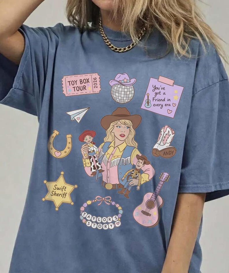 May include: A blue t-shirt featuring a graphic print of a person in a cowboy hat, surrounded by illustrations including a Toy Box Tour ticket, a disco ball, a note saying You've got a friend in every era, a horseshoe, a boot, a Swift Sheriff star, and a guitar.