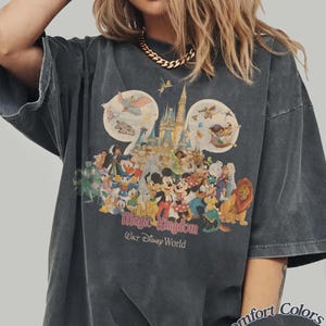 May include: Dark gray t-shirt featuring a colorful graphic of Disney characters and the Magic Kingdom castle. The design includes Mickey Mouse, Donald Duck, and other beloved characters. The text "Magic Kingdom Walt Disney World" is printed below the graphic.