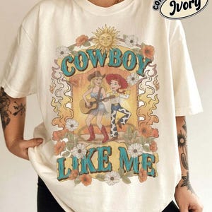 May include: An ivory short-sleeve t-shirt featuring a graphic print with the text "COWBOY LIKE ME" and "COMFORT COLORS IVORY". The design includes a sun, flowers, and two illustrated figures, one playing a guitar and one wearing a cowboy hat.