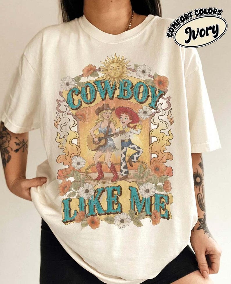 May include: An ivory short-sleeve t-shirt featuring a graphic print with the text "COWBOY LIKE ME" and "COMFORT COLORS IVORY". The design includes a sun, flowers, and two illustrated figures, one playing a guitar and one wearing a cowboy hat.