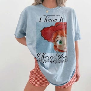 Peut inclure: Un t-shirt bleu clair avec une impression graphique repr&eacute;sentant un personnage de dessin anim&eacute; aux cheveux roux portant un chapeau rouge. Le texte "EST 2026 I Knew It I Knew You" est imprim&eacute; avec une port&eacute;e musicale et des notes noires.