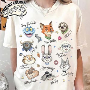 May include: Ivory-colored t-shirt featuring cartoon character illustrations from Zootopia, including Nick Wilde, Judy Hopps, and Flash. The shirt has the words "COMFORT COLORS" and "Ivory" printed on it.