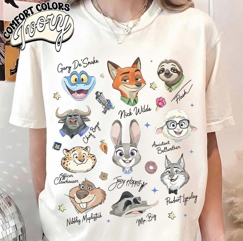 May include: Ivory-colored t-shirt featuring cartoon character illustrations from Zootopia, including Nick Wilde, Judy Hopps, and Flash. The shirt has the words "COMFORT COLORS" and "Ivory" printed on it.