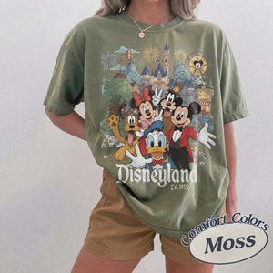 Pu&ograve; includere: Una t-shirt a maniche corte color verde muschio con una stampa grafica di Topolino, Minnie, Paperino, Pippo e Pluto davanti a un castello. Il testo "Disneyland Est. 1955" &egrave; stampato sotto i personaggi con un carattere serif bianco.