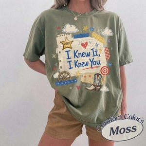 Op de afbeelding: Een olijfgroen t-shirt met korte mouwen en een grafische print van een gescheurd stuk papier met de tekst I Knew It, I Knew You in het blauw. Het ontwerp bevat een gouden sheriff-ster, een rood hart, een wagenwiel, een cowboylaars en sterren.