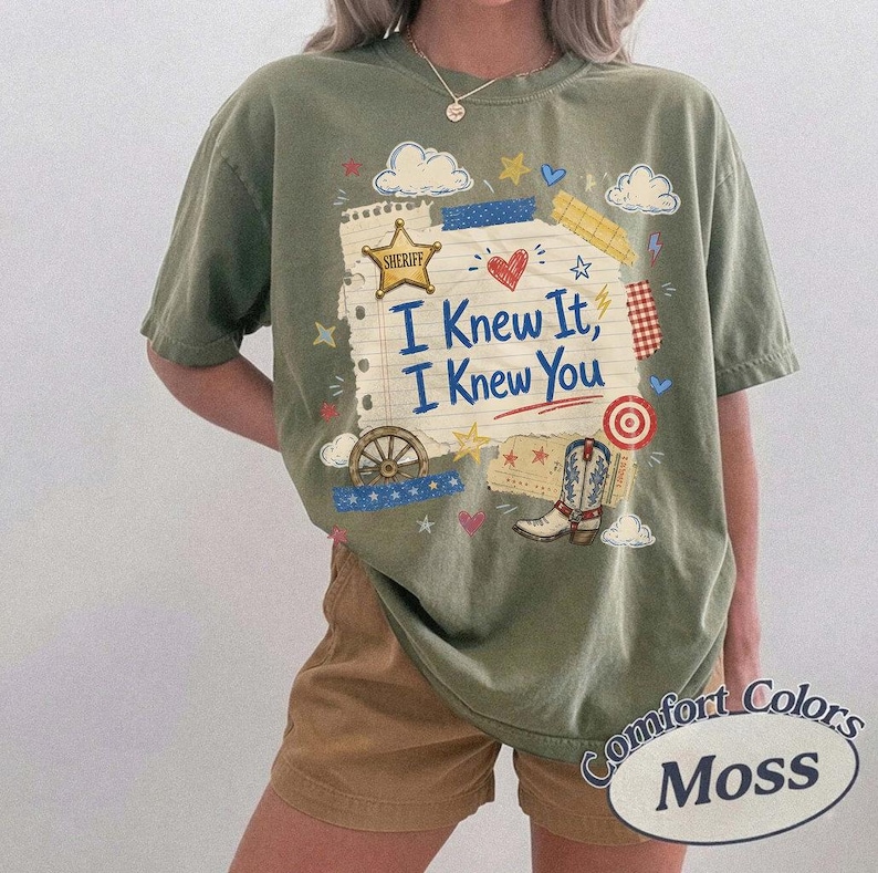 Op de afbeelding: Een olijfgroen t-shirt met korte mouwen en een grafische print van een gescheurd stuk papier met de tekst I Knew It, I Knew You in het blauw. Het ontwerp bevat een gouden sheriff-ster, een rood hart, een wagenwiel, een cowboylaars en sterren.