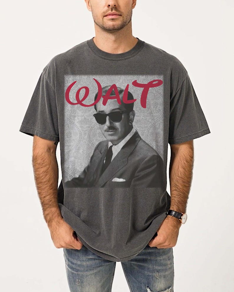 May include: Dark gray t-shirt featuring a black and white image of a man in sunglasses and a suit. The word "WALT" is printed in red cursive above the image. The shirt is a vintage style.