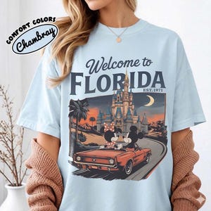 May include: Light blue t-shirt with a graphic that says "Welcome to Florida" and features Mickey and Minnie Mouse in a red convertible in front of a castle. The shirt also has the words "Comfort Colors" and "Chambray" printed on it.