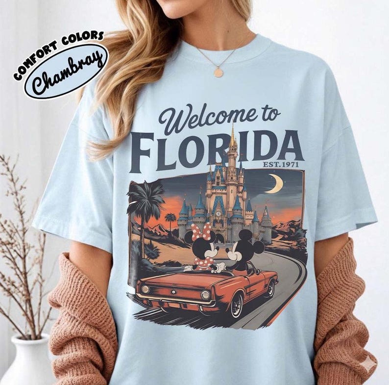 May include: Light blue t-shirt with a graphic that says "Welcome to Florida" and features Mickey and Minnie Mouse in a red convertible in front of a castle. The shirt also has the words "Comfort Colors" and "Chambray" printed on it.