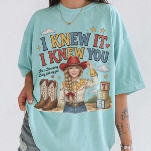 May include: A light blue t-shirt featuring a graphic print of a person in a red cowboy hat, a yellow and white shirt, and blue jeans. The text reads I KNEW IT I KNEW YOU and It's a Love story Baby just say yes. Includes cowboy boots and blocks.