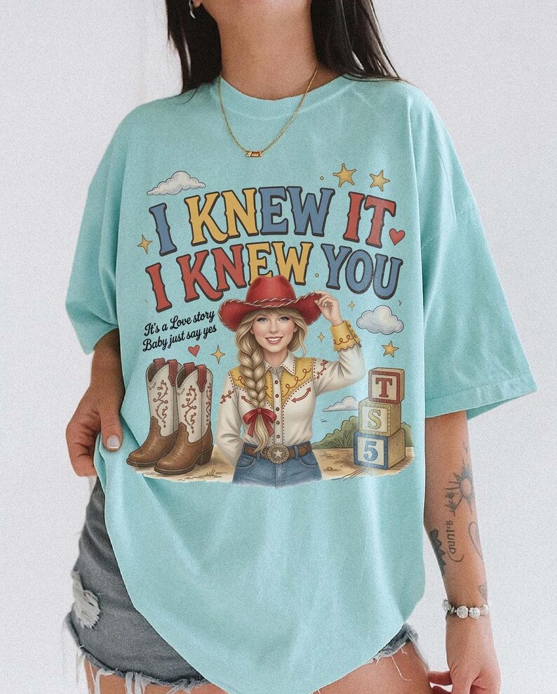 May include: A light blue t-shirt featuring a graphic print of a person in a red cowboy hat, a yellow and white shirt, and blue jeans. The text reads I KNEW IT I KNEW YOU and It's a Love story Baby just say yes. Includes cowboy boots and blocks.