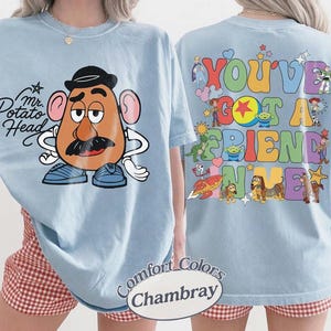 May include: A light blue t-shirt featuring Mr. Potato Head on the front with the text "Mr. Potato Head". The back displays various Toy Story characters and the colorful text "YOU'VE GOT A FRIEND IN ME". The shirt is labeled as Comfort Colors Chambray.