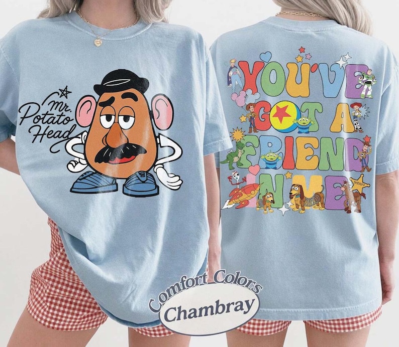 May include: A light blue t-shirt featuring Mr. Potato Head on the front with the text "Mr. Potato Head". The back displays various Toy Story characters and the colorful text "YOU'VE GOT A FRIEND IN ME". The shirt is labeled as Comfort Colors Chambray.