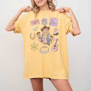 May include: A yellow t-shirt featuring a graphic print of a person in a cowboy hat holding two dolls, surrounded by illustrations including a disco ball, a guitar, a horseshoe, a sheriff badge labeled Swift Sheriff, and text reading Toy Box Tour.