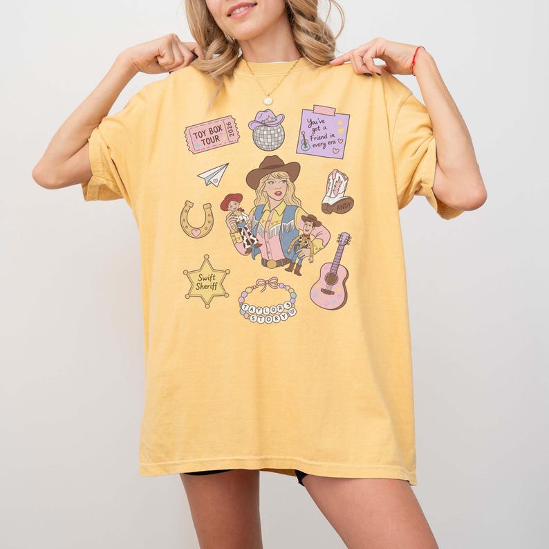 May include: A yellow t-shirt featuring a graphic print of a person in a cowboy hat holding two dolls, surrounded by illustrations including a disco ball, a guitar, a horseshoe, a sheriff badge labeled Swift Sheriff, and text reading Toy Box Tour.