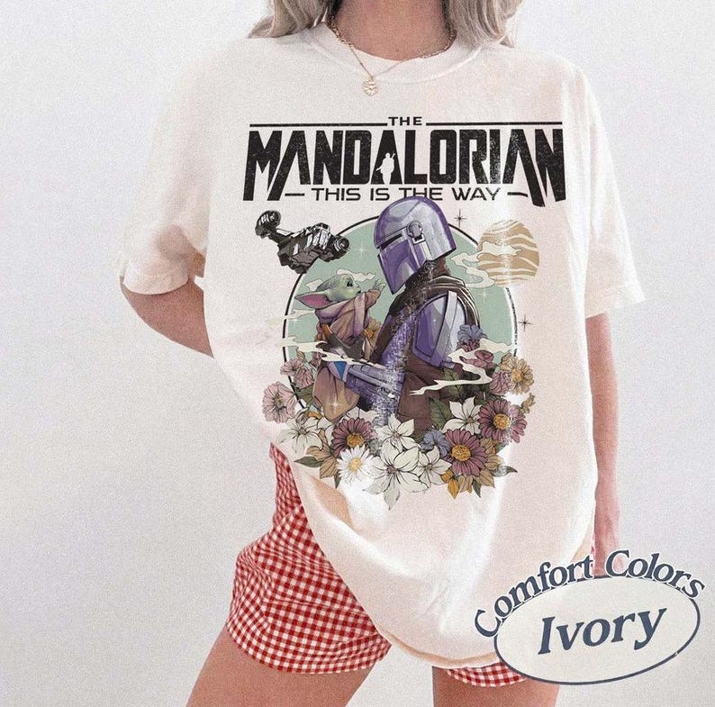 May include: Ivory-colored t-shirt featuring a graphic of The Mandalorian and Grogu surrounded by flowers. The text "THE MANDALORIAN - THIS IS THE WAY" is printed above the image. The shirt is a Comfort Colors brand.