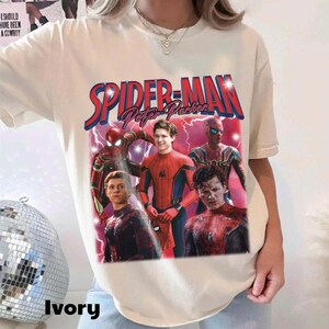 May include: Ivory-colored t-shirt featuring a graphic of Spider-Man with the text "SPIDER-MAN Peter Parker" in red and blue. The design includes multiple images of Spider-Man in various suits and poses, with lightning effects.
