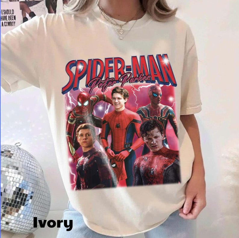 May include: Ivory-colored t-shirt featuring a graphic of Spider-Man with the text "SPIDER-MAN Peter Parker" in red and blue. The design includes multiple images of Spider-Man in various suits and poses, with lightning effects.