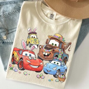 Pode incluir: Camiseta creme com um gr&aacute;fico colorido de carros animados decorados para a P&aacute;scoa. O design inclui Rel&acirc;mpago McQueen, Mate e outros personagens com ovos de P&aacute;scoa e orelhas de coelho. Um chap&eacute;u bege e jeans azuis est&atilde;o no fundo.