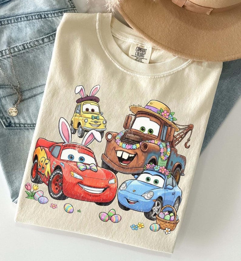 Pode incluir: Camiseta creme com um gr&aacute;fico colorido de carros animados decorados para a P&aacute;scoa. O design inclui Rel&acirc;mpago McQueen, Mate e outros personagens com ovos de P&aacute;scoa e orelhas de coelho. Um chap&eacute;u bege e jeans azuis est&atilde;o no fundo.