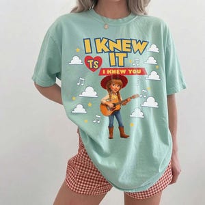 May include: A light green t-shirt featuring a graphic of a cowgirl playing an acoustic guitar. The text reads I KNEW IT and I KNEW YOU, accompanied by a red heart, yellow stars, and white clouds. The design includes musical note symbols.