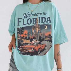 May include: A light blue t-shirt with the text "Welcome to Florida" and a graphic of Mickey and Minnie Mouse in a red convertible in front of a castle. The shirt has short sleeves.