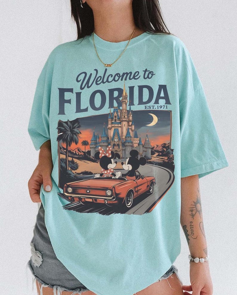 May include: A light blue t-shirt with the text "Welcome to Florida" and a graphic of Mickey and Minnie Mouse in a red convertible in front of a castle. The shirt has short sleeves.
