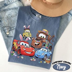 Pode incluir: Camiseta Comfort Colors azul marinho com carros de desenho animado com orelhas de coelhinho da P&aacute;scoa e decora&ccedil;&otilde;es florais. O design inclui Rel&acirc;mpago McQueen, Mate e outros personagens, junto com ovos de P&aacute;scoa e uma cesta.