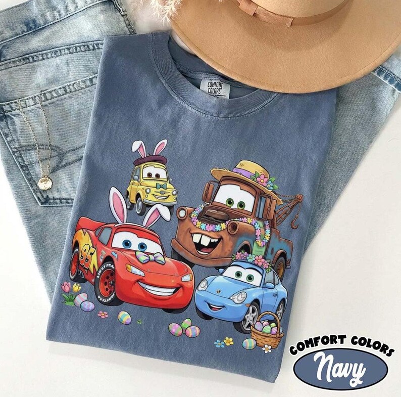 Pode incluir: Camiseta Comfort Colors azul marinho com carros de desenho animado com orelhas de coelhinho da P&aacute;scoa e decora&ccedil;&otilde;es florais. O design inclui Rel&acirc;mpago McQueen, Mate e outros personagens, junto com ovos de P&aacute;scoa e uma cesta.