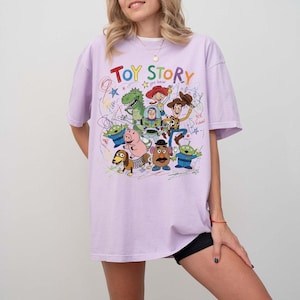 May include: A light purple t-shirt featuring a colorful graphic of characters from the animated film "Toy Story." The design includes the words "Toy Story" in a playful font, along with drawings of Woody, Buzz Lightyear, and other characters.
