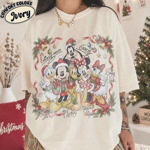 Puede incluir: Una camiseta color marfil con un gráfico festivo de Mickey Mouse, Minnie Mouse, Donald Duck, Daisy Duck, Goofy y Pluto con ropa navideña. El diseño incluye ramas de pino, cintas, campanas y los nombres de los personajes escritos a mano.