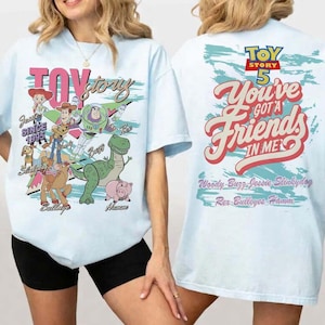 Pode incluir: Uma t-shirt azul-clara com gr&aacute;ficos de Toy Story. A frente exibe Woody, Buzz Lightyear, Jessie, Slinky Dog, Bullseye e Hamm com o texto "Toy Story SINCE 1995". As costas apresentam o texto "TOY STORY 5 You've Got A Friends In Me".