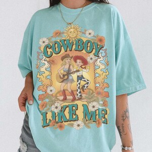 May include: A light blue t-shirt featuring a graphic print with the text "COWBOY LIKE ME" in large blue letters. The design includes a sun, flowers, and two figures, one playing a guitar and the other wearing a cowboy hat, surrounded by flames.